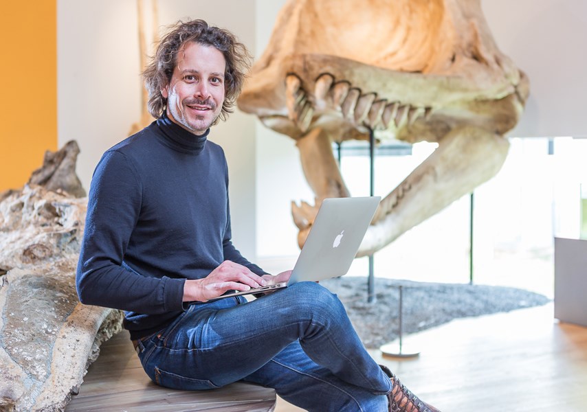 Man op laptop in museum, dinosaurus schedel op de achtergrond