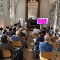 Foto achterin de Van Assendelftkapel gericht op het podium en publiek, de zaal waar de bijeenkomst gehouden werd.