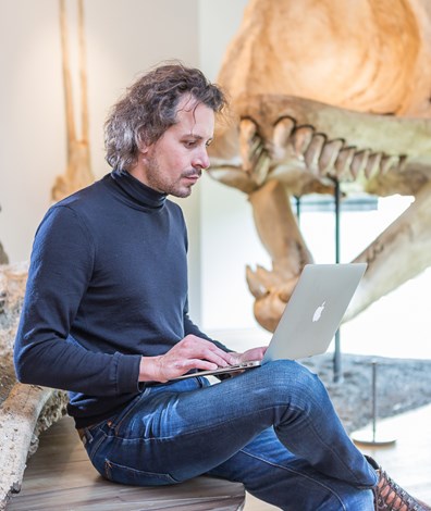 Man zit met laptop op schoot met op achtergrond skelet van dinosaurus