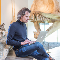 Man zit met laptop op schoot met op achtergrond skelet van dinosaurus